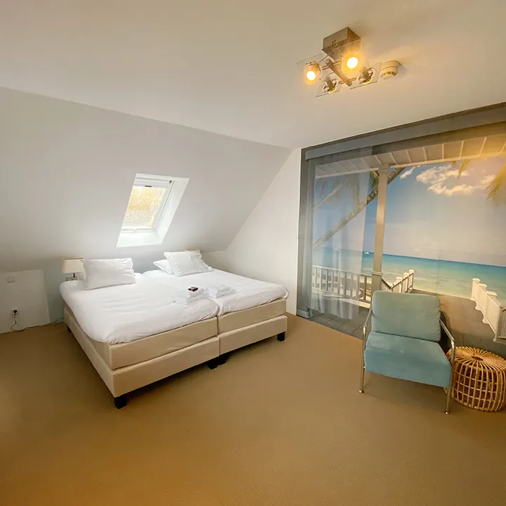 Een bed onder een schuin dak met dakraam, met een groot strandtafereel op de wand.