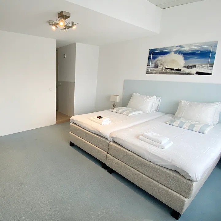 Een lichte kamer met lichtblauwe accenten en een ingelijst zeegezicht boven het bed.