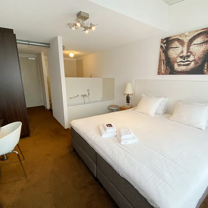 Een groot Boeddhahoofd op canvas boven het bed, met een bad achter een lage witte muur en okerkleurig tapijt.