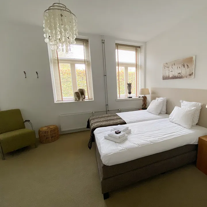 Een schelpenluchter bij het raam, een olijfgroene fauteuil en twee bedden onder een ingelijste prent.