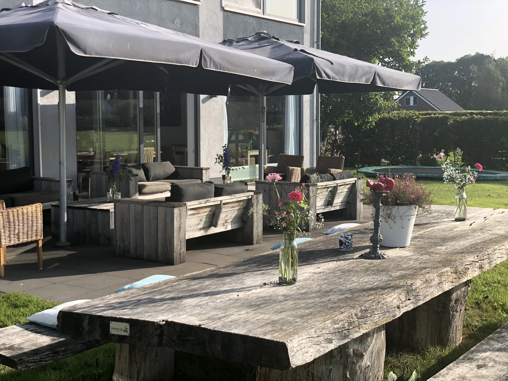 Het terras: een lange verweerde houten tafel met banken, parasols open, bloemen in vazen en de glazen pui van het huis erachter.